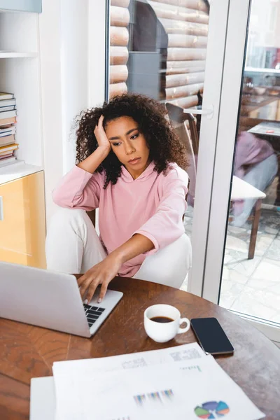 Selective focus of african american freelancer looking at laptop near smartphone with blank screen, charts and graphs — Stock Photo