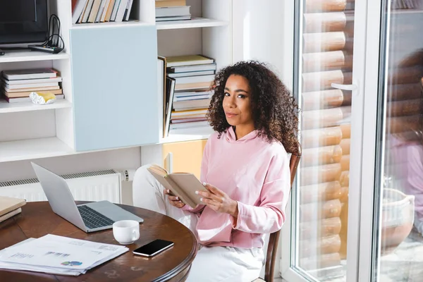 Cheerful african american freelancer reading book near gadgets and cup — Stock Photo