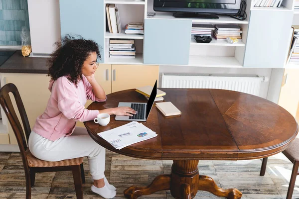 Curly african american freelancer using laptop near cup, books and documents — Stock Photo