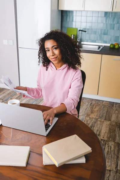 Selective focus of happy african american freelancer using laptop and holding documents near cup and books — Stock Photo