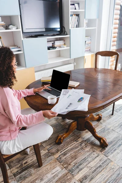 African american freelancer using laptop with blank screen and holding documents near cup and books — Stock Photo