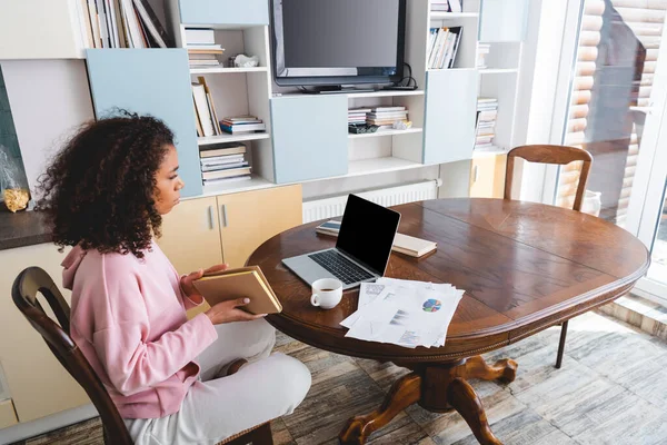 Afroamericano freelancer celebración de libro cerca de la computadora portátil con pantalla en blanco, taza y documentos - foto de stock