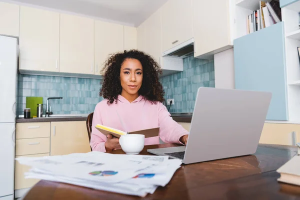 Selektiver Fokus des lockigen afrikanisch-amerikanischen Freelancers, der auf die Kamera in der Nähe von Laptop, Tasse, Diagrammen und Grafiken blickt — Stockfoto