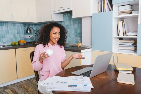 Feliz africano americano chica gesto mientras sostiene taza y tener video chat en casa - foto de stock