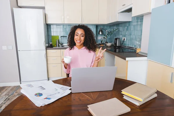 Glückliches afrikanisch-amerikanisches Mädchen zeigt Friedenszeichen, während es Tasse in der Hand hält und zu Hause einen Videochat führt — Stockfoto