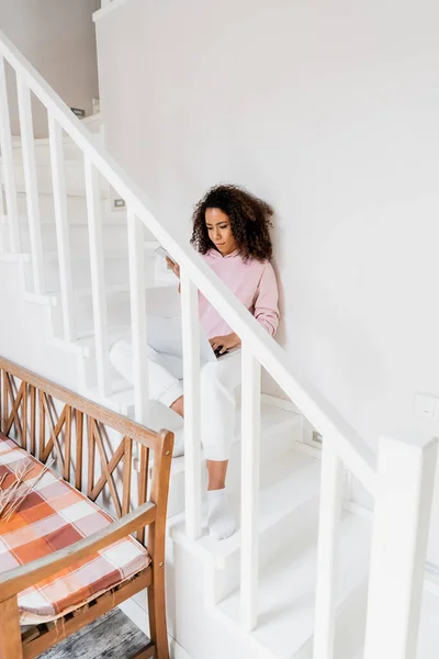 Freelancer afroamericano sentado en las escaleras mientras usa el portátil y sostiene el teléfono inteligente - foto de stock