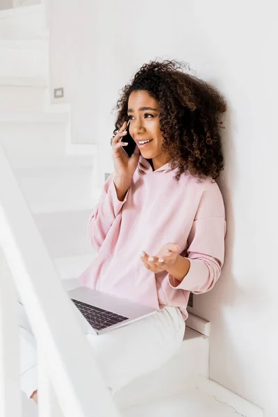 Freelancer afro-americano feliz sentado em escadas enquanto usa laptop e falando no smartphone — Fotografia de Stock