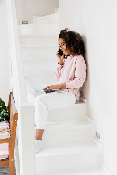 Fröhliche afrikanisch-amerikanische Freiberuflerin sitzt auf der Treppe, während sie Laptop benutzt und mit dem Smartphone spricht — Stockfoto