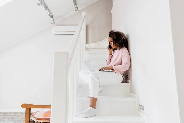 Displeased african american freelancer sitting on stairs while using laptop and talking on smartphone — Stock Photo