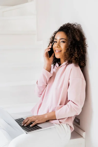 Smiling african american freelancer sitting on stairs while using laptop and talking on smartphone — Stock Photo