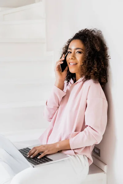 Souriant afro-américain freelance assis sur les escaliers tout en utilisant un ordinateur portable et parler sur smartphone — Photo de stock