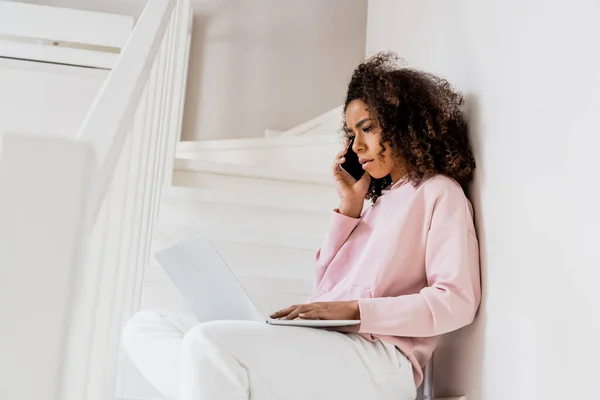 Upset african american freelancer sitting on stairs while using laptop and talking on smartphone — Stock Photo