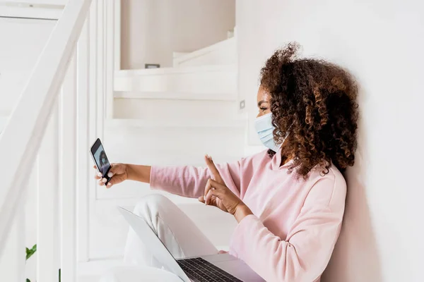 Encaracolado Africano americano freelancer em máscara médica sentado em escadas e mostrando sinal de paz ao tomar selfie perto laptop — Fotografia de Stock