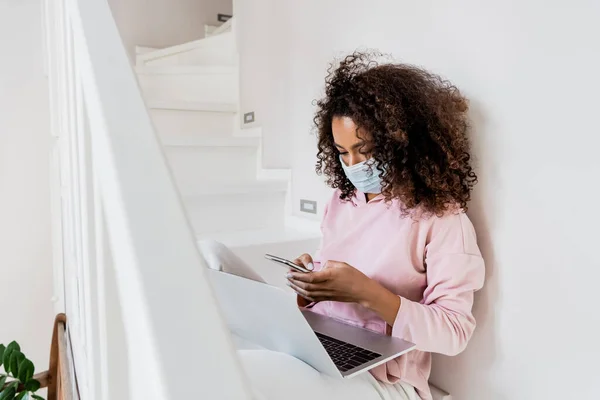 Selective focus of curly african american freelancer in medical mask sitting on stairs and using smartphone — Stock Photo
