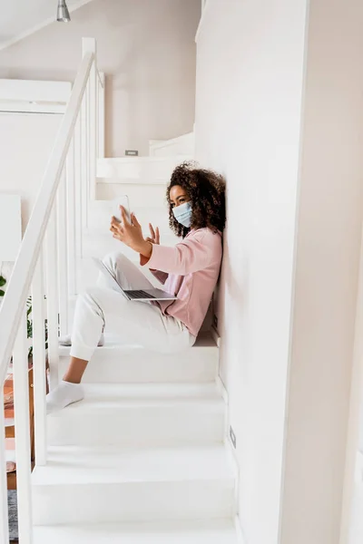 Afroamericano freelancer en máscara médica sentado en escaleras y mostrando señal de paz mientras toma selfie cerca de laptop - foto de stock