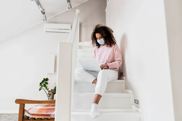 Молодой африканский американский фрилансер в медицинской маске сидит на лестнице и использует ноутбук — стоковое фото