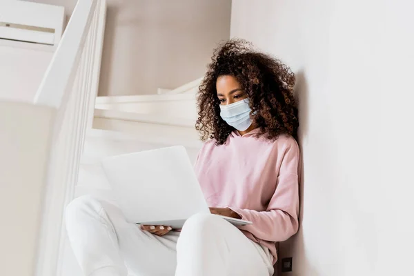 Encaracolado Africano americano freelancer em máscara médica sentado em escadas e usando laptop — Fotografia de Stock