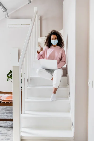 Curly african american freelancer in medical mask sitting on stairs and showing thumb up near laptop — Stock Photo