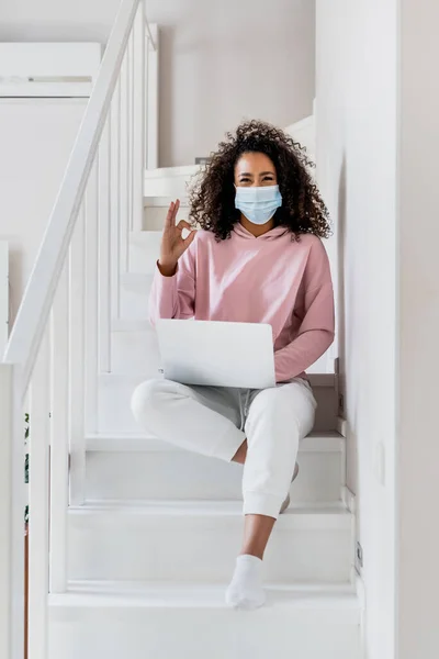 Curly african american freelancer in medical mask sitting on stairs and showing ok sign near laptop — Stock Photo