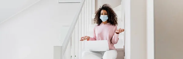 Concept panoramique de freelance afro-américain confus dans le masque médical assis sur les escaliers et montrant geste haussant les épaules près d'un ordinateur portable — Photo de stock
