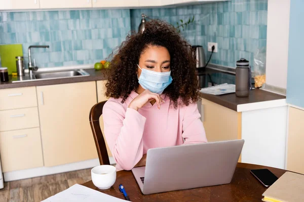 Encaracolado Africano americano freelancer em máscara médica olhando para laptop — Fotografia de Stock