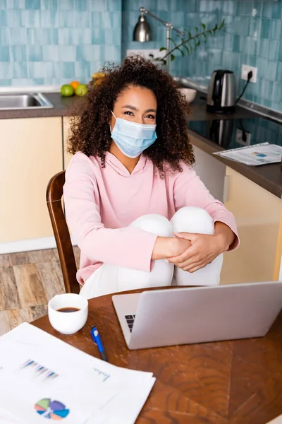 Freelancer americano africano encaracolado em máscara médica sentado perto de laptop, copo, gráficos e gráficos — Fotografia de Stock