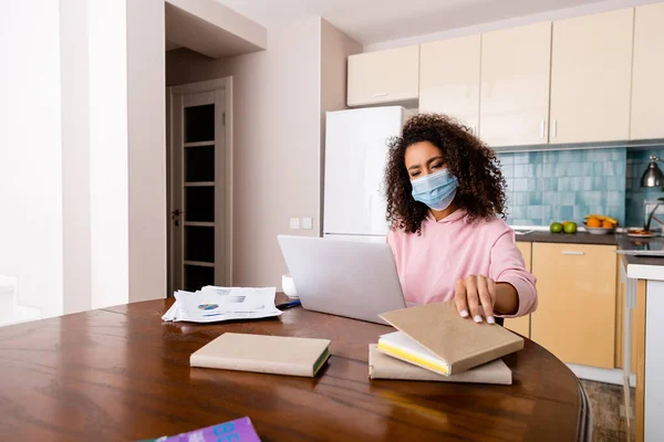 Encaracolado Africano americano freelancer em máscara médica tocando livro perto de laptop — Fotografia de Stock