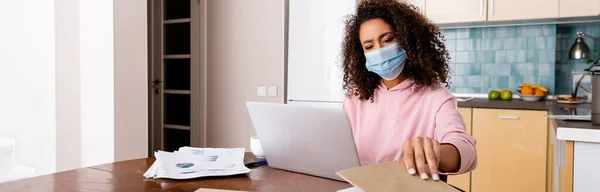 Concepto horizontal de rizado afroamericano freelancer en máscara médica tocando libro cerca del ordenador portátil - foto de stock