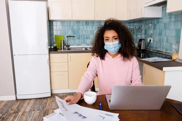Curly african american freelancer in medical mask touching charts and graphs near laptop — Stock Photo