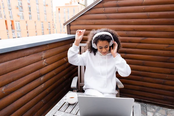 Feliz afroamericano freelancer en auriculares inalámbricos escuchando música cerca de la computadora portátil y taza de café - foto de stock