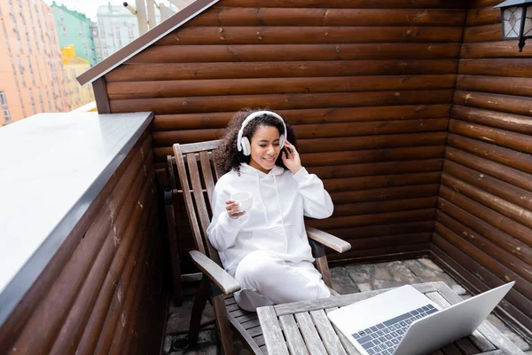 Cheerful african american freelancer in wireless headphones listening music near laptop and holding cup of coffee — Stock Photo