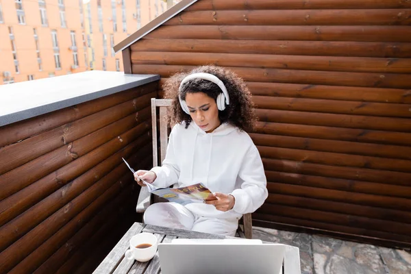 Curly african american freelancer in wireless headphones reading magazine near laptop and cup of coffee — Stock Photo