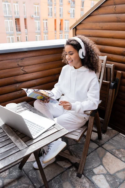 Feliz afroamericano freelancer en auriculares inalámbricos revista de lectura cerca de portátil y taza - foto de stock