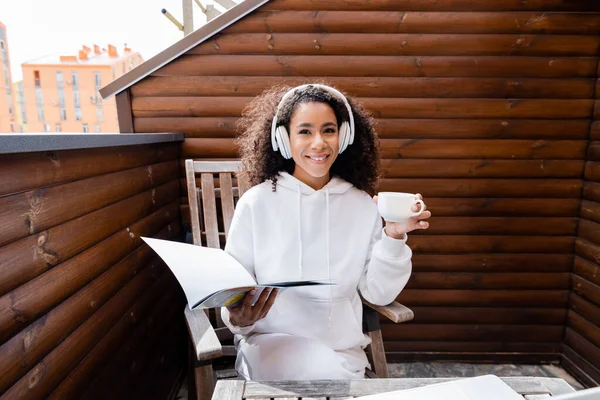 Glückliches afrikanisch-amerikanisches Mädchen in drahtlosen Kopfhörern mit Magazin und Tasse — Stockfoto