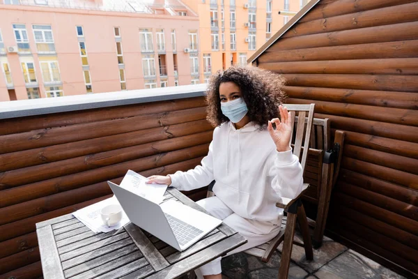 Chica afroamericana en máscara médica mostrando ok signo cerca de portátil, taza, gráficos y gráficos - foto de stock