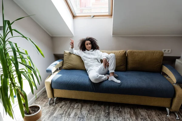 Menina americana africana alegre mostrando sinal de paz enquanto sentado no sofá em casa — Fotografia de Stock