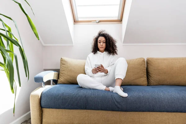 Chica americana africana rizada usando teléfono inteligente y sentado en el sofá en casa - foto de stock