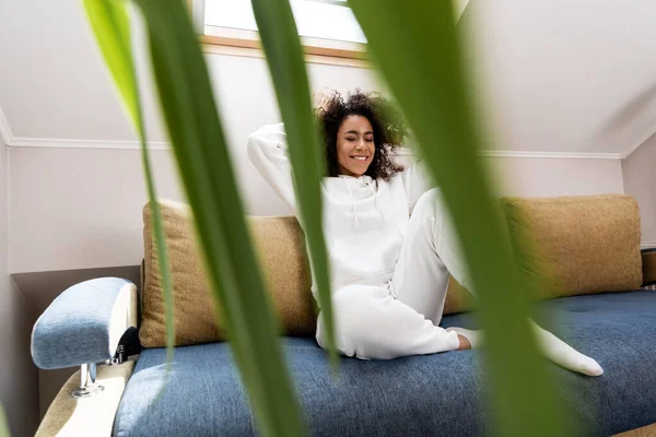 Selektiver Fokus einer glücklichen Afroamerikanerin, die auf einem Sofa in der Nähe einer Pflanze sitzt — Stockfoto