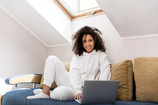Alegre afroamericano freelancer sentado en el sofá y el uso de la computadora portátil en casa — Stock Photo