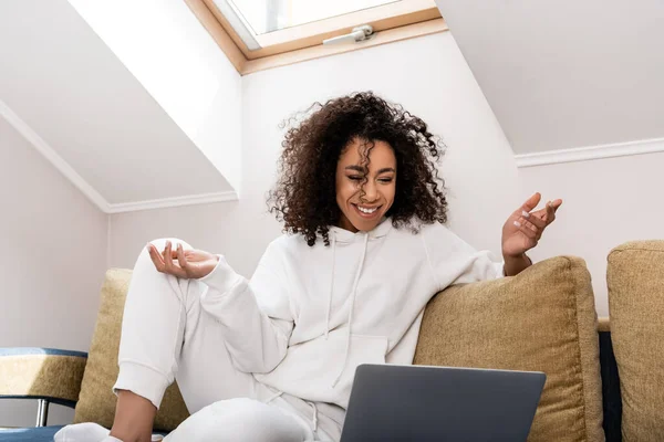 Happy african american freelancer sitting on sofa and gesturing near laptop — Stock Photo