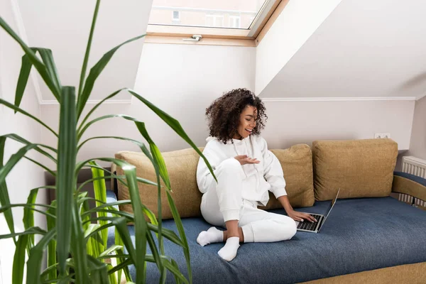 Foyer sélectif de fille afro-américaine heureuse assise sur le canapé et gestuelle près d'un ordinateur portable tout en ayant un appel vidéo — Photo de stock