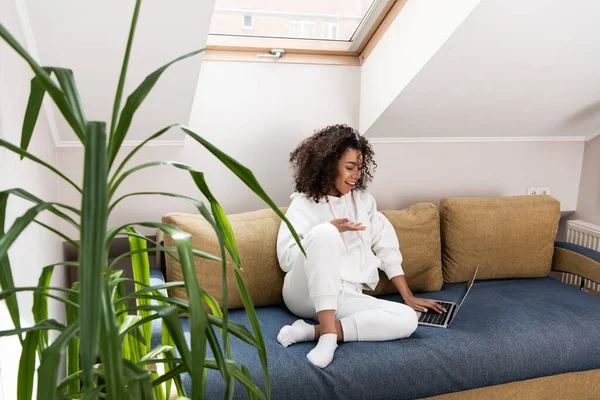 Messa a fuoco selettiva di felice ragazza afro-americana seduta sul divano e gesticolare vicino al computer portatile mentre ha videochiamata — Foto stock