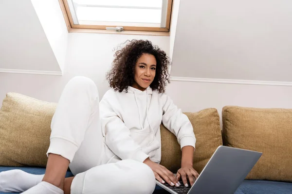 Feliz afroamericano freelancer sentado en el sofá y utilizando el ordenador portátil - foto de stock