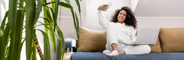 Tienda panorámica de feliz joven afroamericano freelancer tomando selfie cerca del ordenador portátil - foto de stock