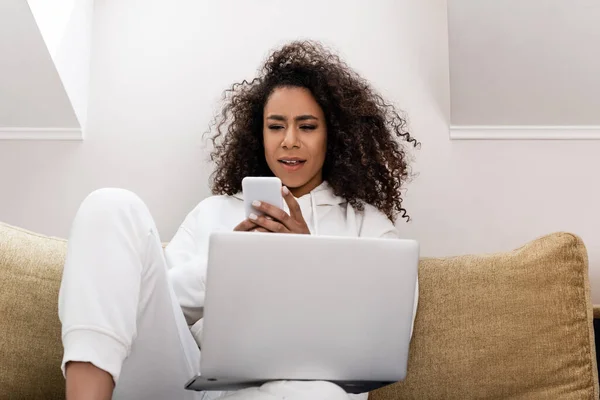 Joven afroamericano freelancer utilizando teléfono inteligente cerca del ordenador portátil - foto de stock