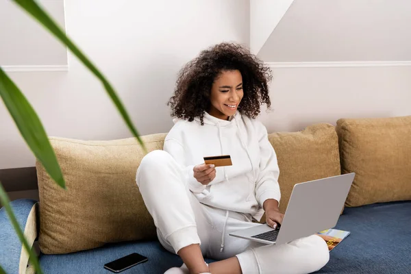 Enfoque selectivo de la niña afroamericana feliz celebración de la tarjeta de crédito cerca de la computadora portátil mientras está sentado en el sofá - foto de stock