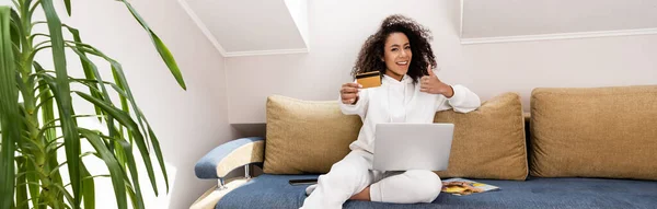 Panoramische Konzept der glücklichen afrikanisch-amerikanischen Mädchen zeigt Daumen hoch und hält Kreditkarte in der Nähe Laptop — Stockfoto