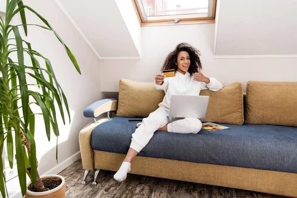 Selective focus of happy african american girl showing thumb up and holding credit card near laptop — Stock Photo