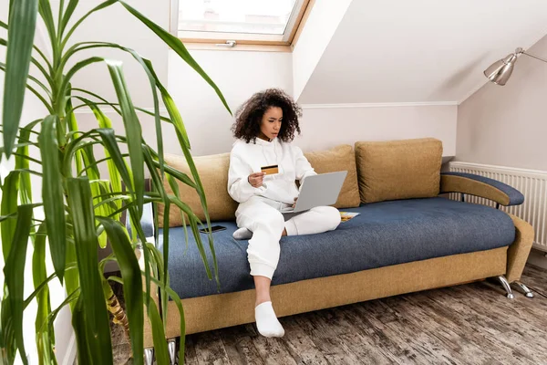 Enfoque selectivo de chica afroamericana sosteniendo la tarjeta de crédito cerca de la computadora portátil mientras está sentado en el sofá - foto de stock