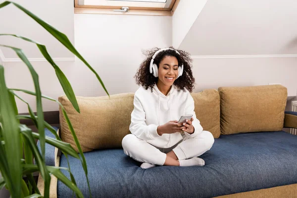 Enfoque selectivo de la chica afroamericana feliz en auriculares inalámbricos usando teléfono inteligente mientras está sentado en el sofá - foto de stock